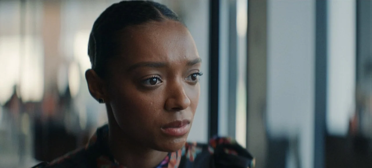 Babygirl (2024) movie still: closeup — A bright indoor office space with a large window in the background. A young woman with dark hair stands near…