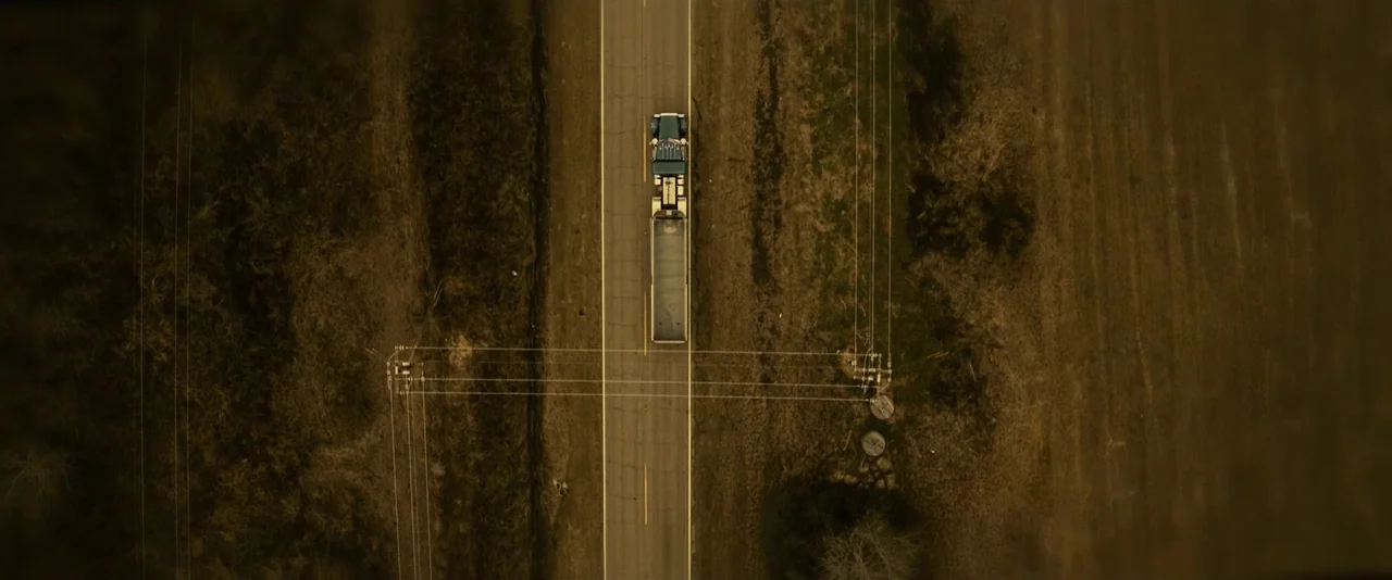 Blaze (2018) movie still: wide — A top-down view shows a semi-truck traveling on a straight asphalt highway. The surrounding landscape consists of…