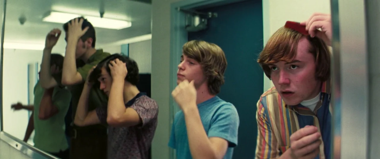 Licorice Pizza (2021) movie still: medium — A public restroom with light-colored walls and a long mirror reflecting several young men. The men stand…