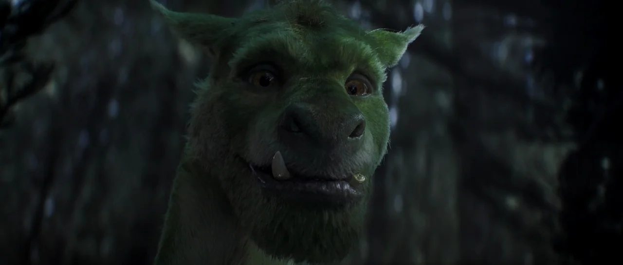 Pete’s Dragon (2016) movie still: closeup — A green furry creature is centered in a dark, out-of-focus woodland setting. The creature looks forward…