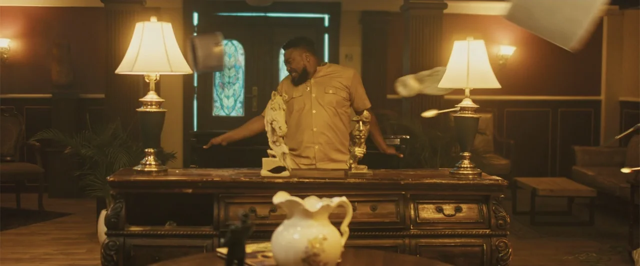 Casa De Novia (2024) movie still: medium — A dimly lit room contains a large dark wood desk with ornate decorative sculptures. A bearded man in a…