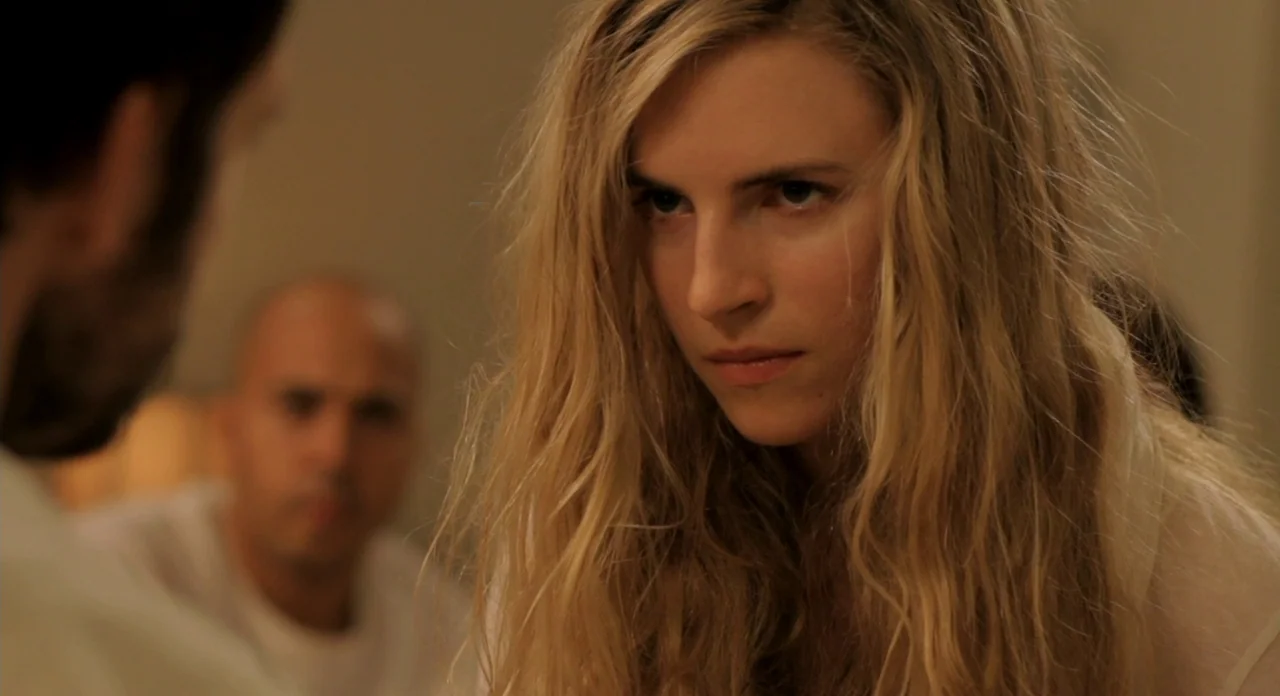 Sound Of My Voice (2011) movie still: closeup — A dimly lit room with blurred figures in the background. A woman with long blonde hair stares ahead…