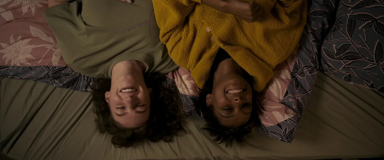 Talk To Me (2022) movie still: medium — Two women lie on a bed with floral-patterned pillows and a green sheet. They are looking directly upward and…