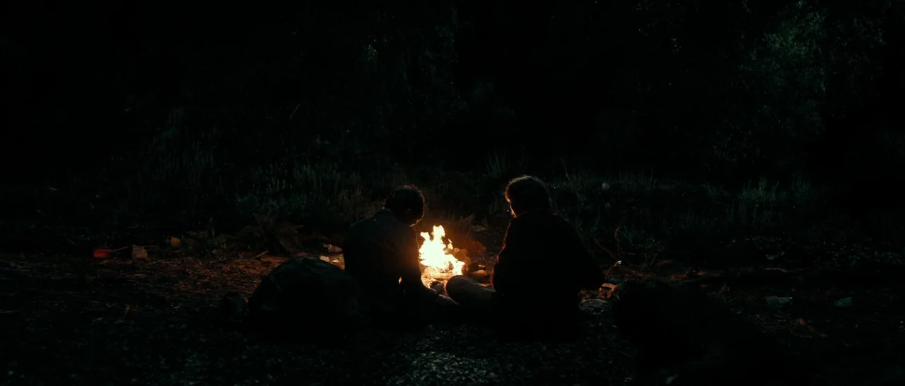 Swiss Army Man (2016) movie still: wide — Two individuals sit on the dark, uneven ground in a remote wooded clearing at night. They are positioned…