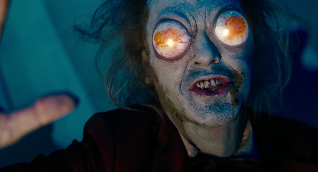 Beetlejuice Beetlejuice (2024) movie still: closeup — A dark environment surrounds a pale male figure with grotesquely protruding, glowing orange…