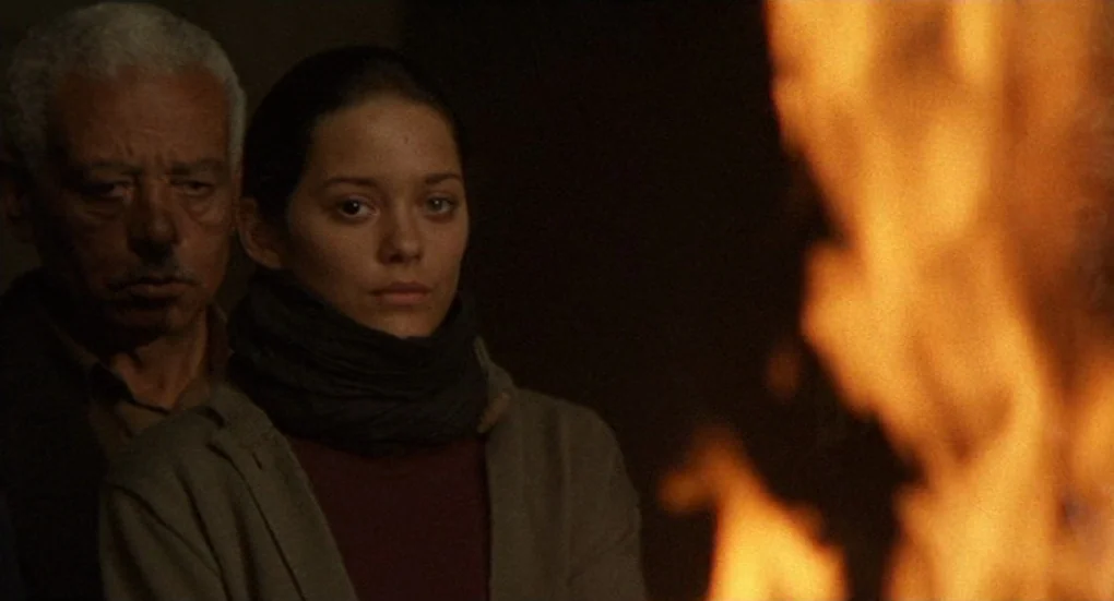 Furia (1999) movie still: medium — A dimly lit room is illuminated only by a nearby flickering fire. A young woman and an older man stand close…