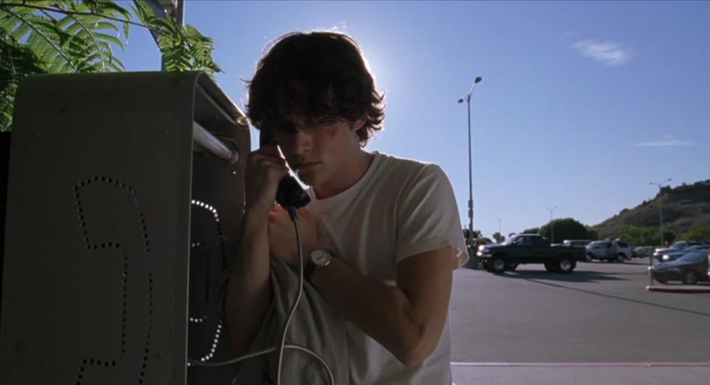 Brick (2005) movie still: medium — An asphalt parking lot with distant hills and parked vehicles under a bright sky. A young man stands at a public…