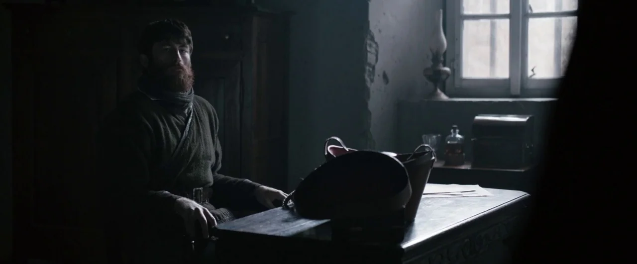 Black ’47 (2018) movie still: medium — A dimly lit room with distressed plaster walls features a single window and wooden furniture. A bearded man…