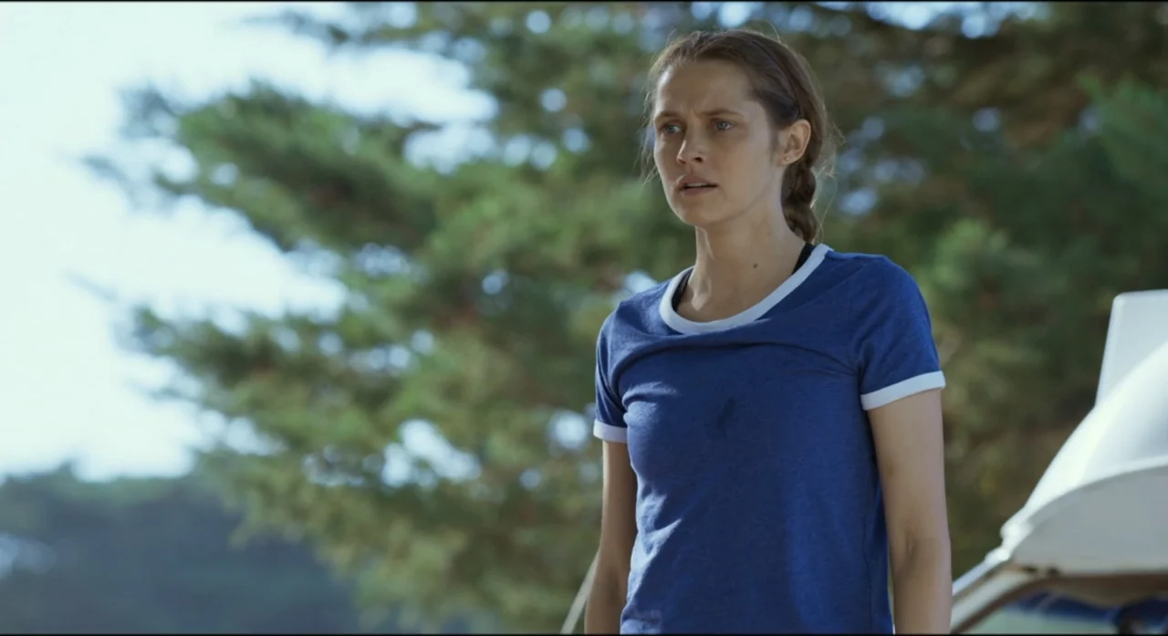 Ride Like a Girl (2019) movie still: medium — A woman stands outdoors in front of out-of-focus pine trees during the day. The woman in a blue…