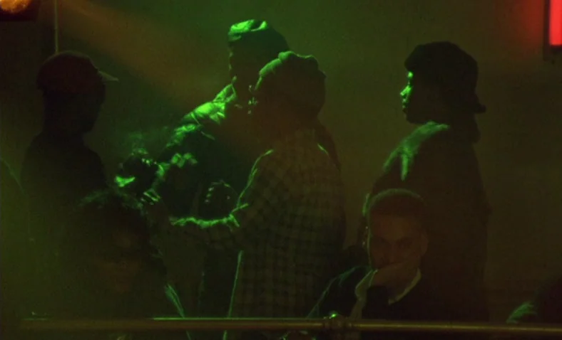 Paris Is Burning (1990) movie still: medium — A dark, smoke-filled room is lit by heavy green atmospheric lighting and neon. A group of people stand…