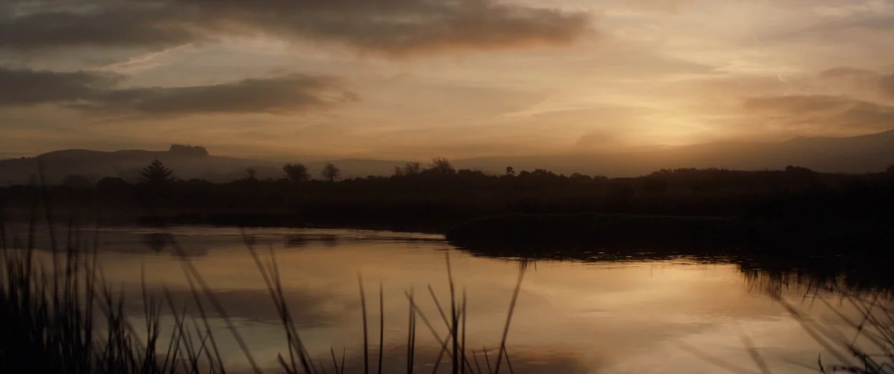 Never Grow Old (2019) movie still: establishing — A tranquil river winds through a misty landscape under a golden twilight sky. Tall grasses frame…