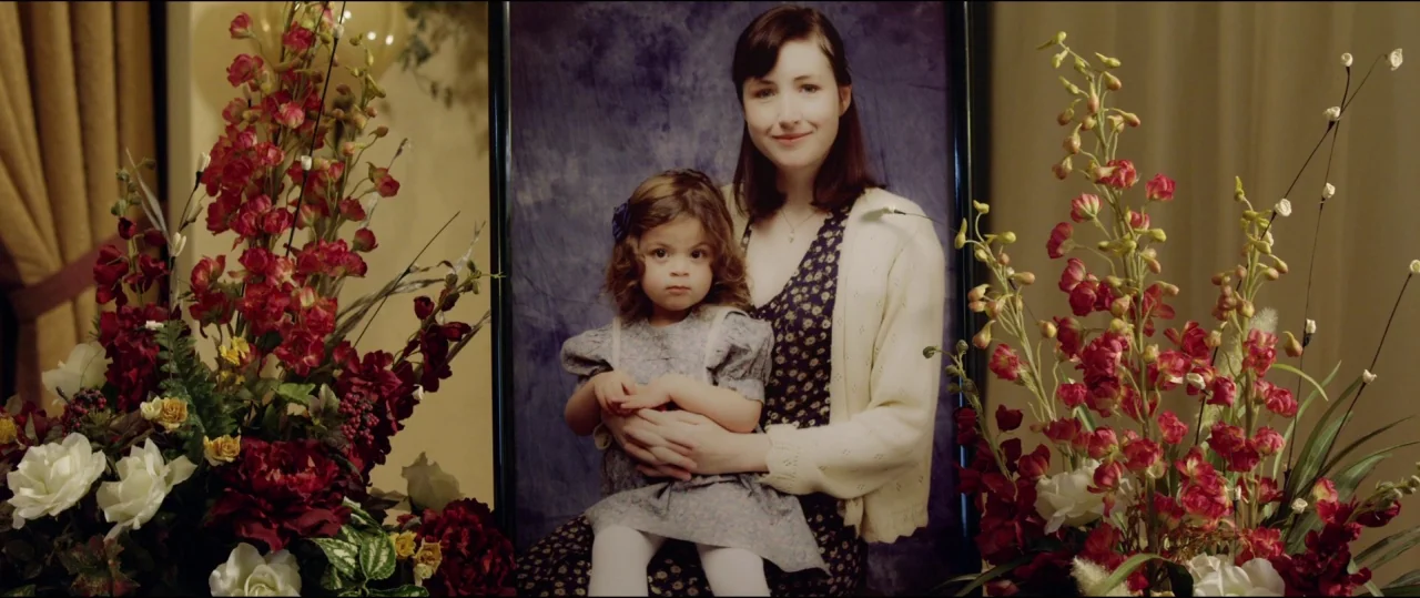 Buster’s Mal Heart (2016) movie still: medium — A studio portrait of a woman and a child positioned before a blue backdrop. Large floral bouquets…