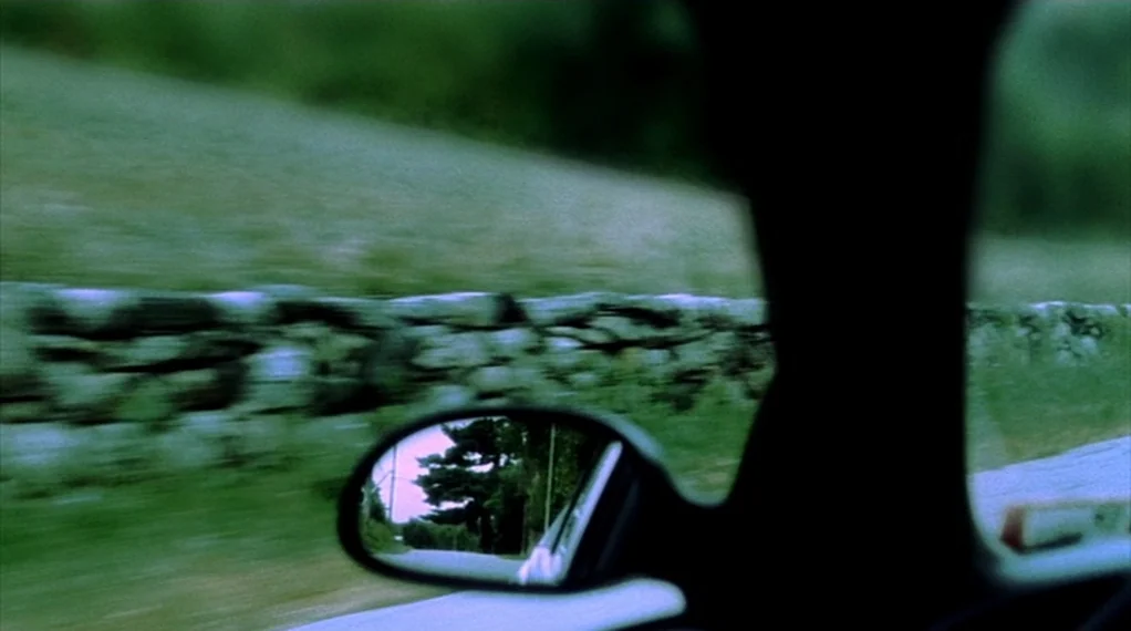 Broken Flowers movie still: medium — A view from inside a moving vehicle shows a stone wall passing by quickly. A side view mirror reflects a rural…