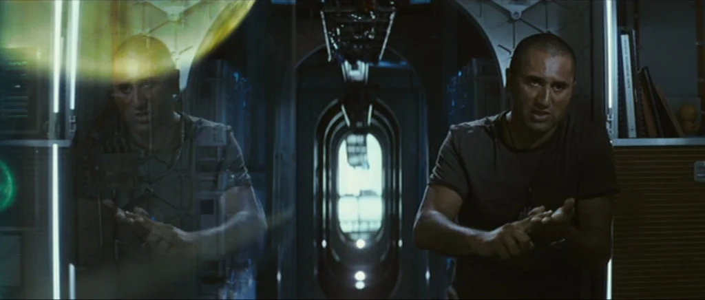 Sunshine movie still: medium — A dark, industrial spaceship hallway with metallic paneling and a glowing rectangular bulkhead opening. A man stands…