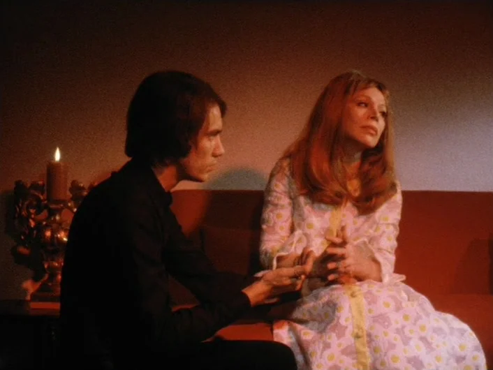 Eika Katappa (1969) movie still: medium — A dimly lit room with an orange sofa against a bare wall. A man sits and reaches toward the woman as she…