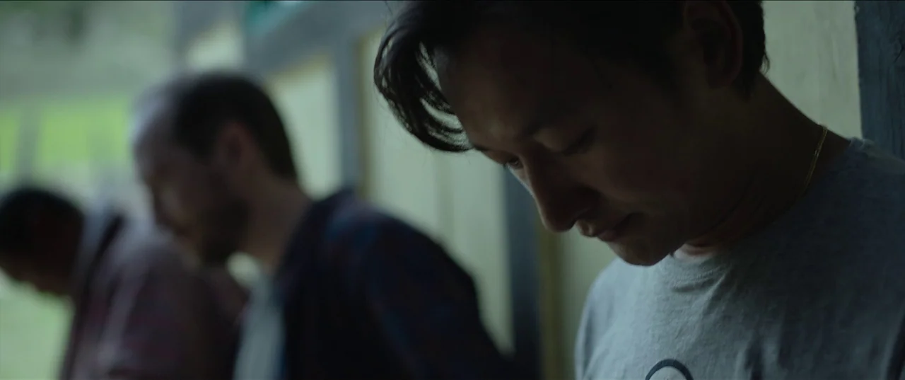 The Monk and The Gun (2023) movie still: closeup — A dimly lit indoor passageway with pale yellow walls and dark vertical structural beams. An East…