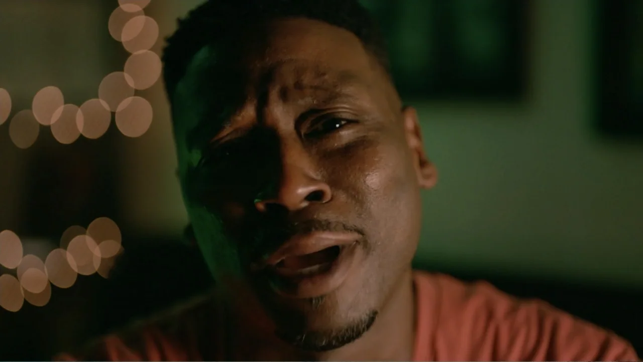 Between the World and Me (2020) movie still: closeup — A dimly lit interior space features out-of-focus fairy lights in the background. A man speaks…