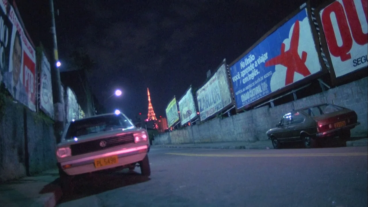 Powaqqatsi (1988) movie still: wide — An empty urban street at night is flanked by rows of large advertising billboards. A tall illuminated…