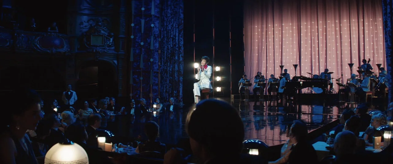 Judy (2019) movie still: wide — A dim supper club featuring an orchestra and audience members sitting at small tables. A lone female singer performs…