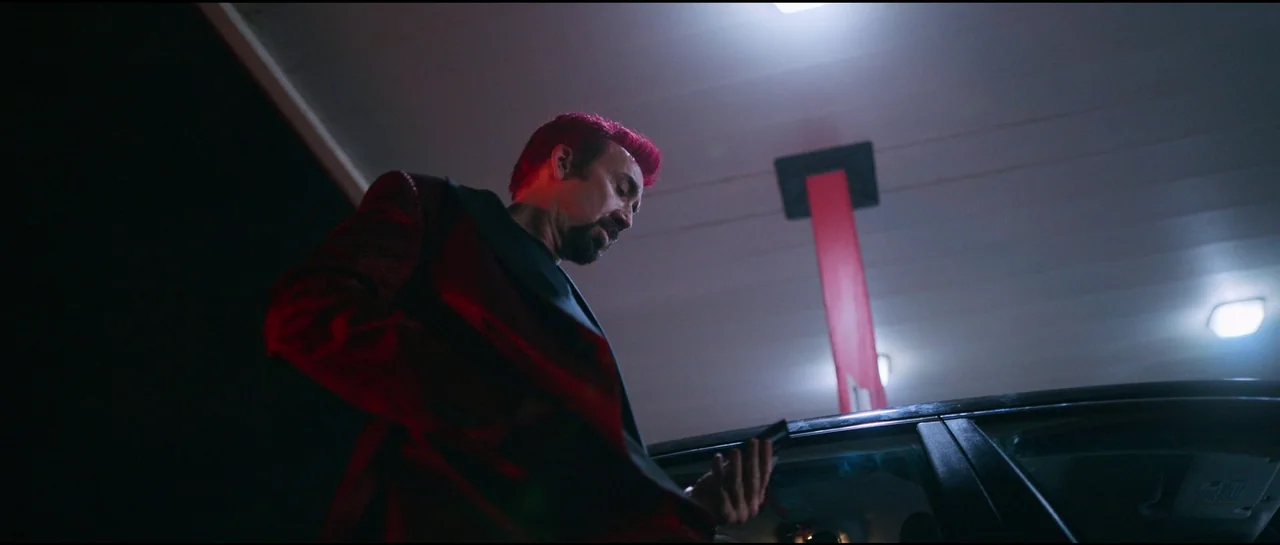 Sympathy For The Devil (2023) movie still: medium — A man stands beneath the metal canopy of a dark, late-night gas station. He looks down at his…