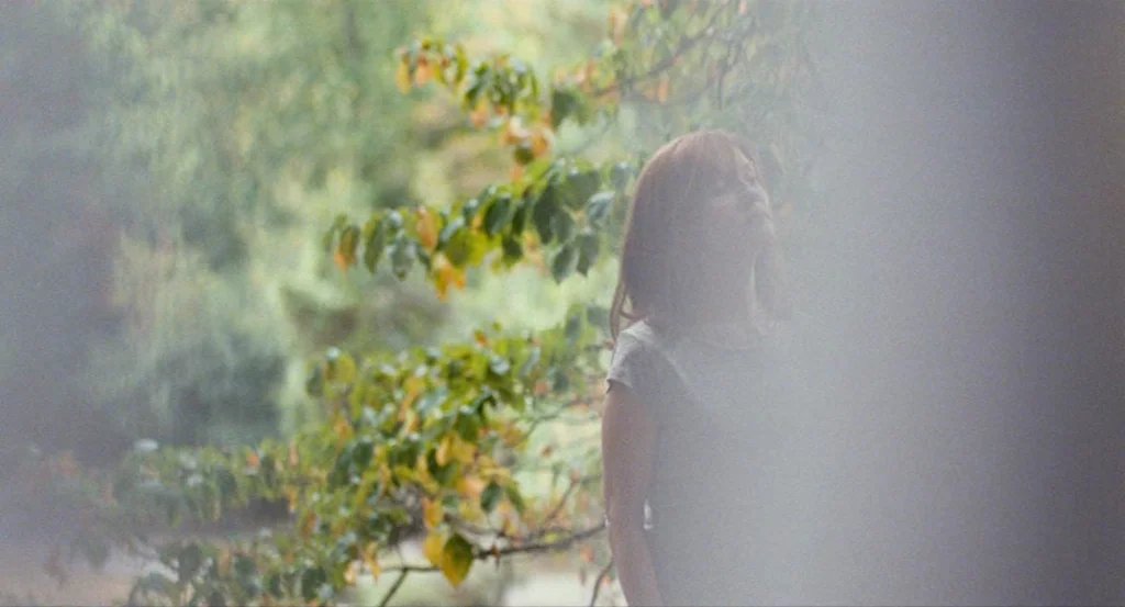 Louder Than Bombs (2015) movie still: medium — A woman stands in an area densely covered with green trees and yellowing leaves. The subject gazes…