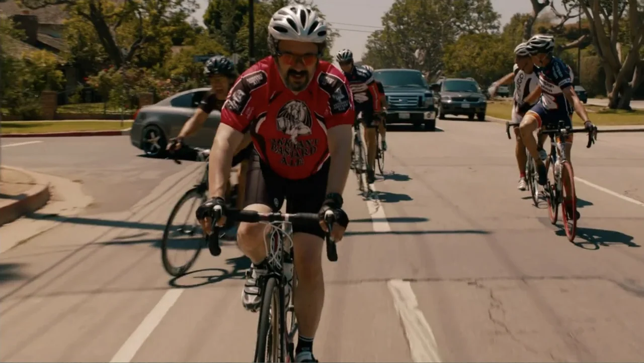 This Is 40 (2012) movie still: medium — A residential street lined with mature trees and parked cars in the background. A group of cyclists rides…