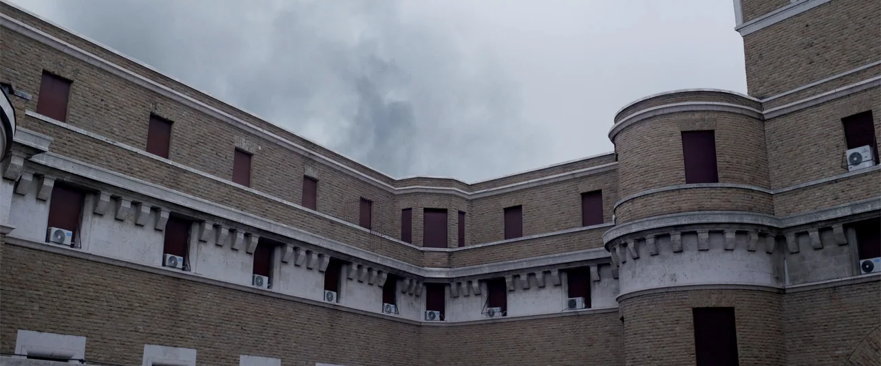 Conclave (2024) movie still: establishing — A stone building with multiple levels features dark window shutters and exterior wall units. The facade…