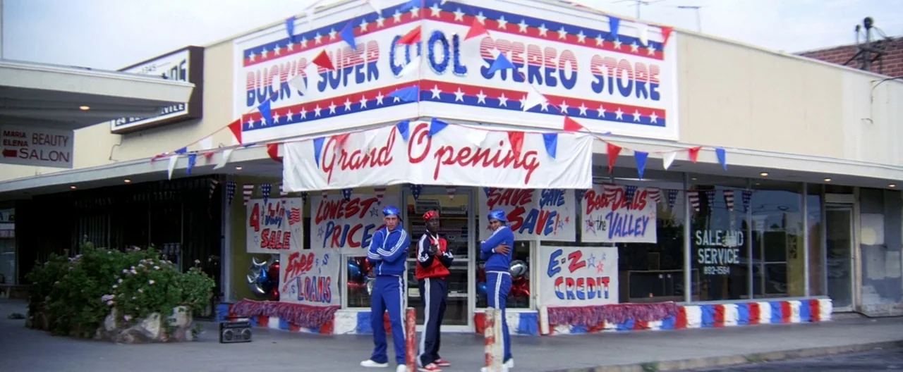 Boogie Nights movie still: wide — A retail strip mall storefront displays patriotic red, white, and blue banners and signage. Three men in track…