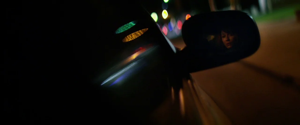 Lost River (2014) movie still: closeup — The exterior of a dark vehicle is visible at night under street lighting. A female face is reflected in the…