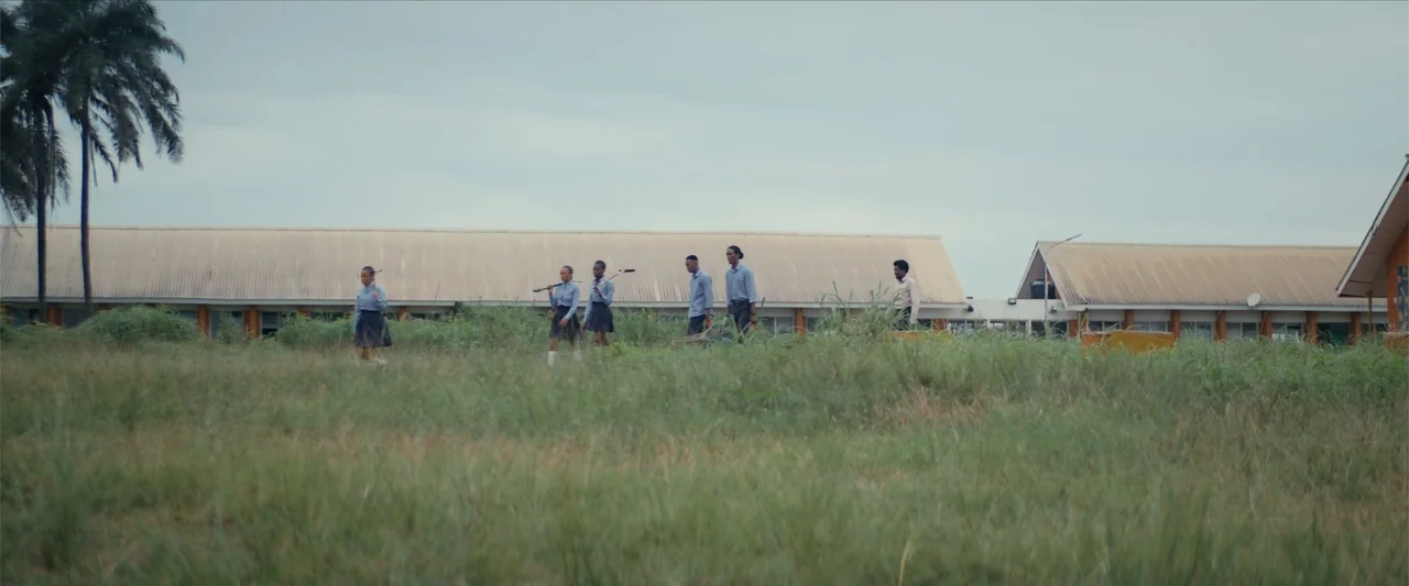 Ms. Kanyin (2025) movie still: wide — A grassy schoolyard sits in front of long, low-slung, single-story institutional buildings. A group of five…