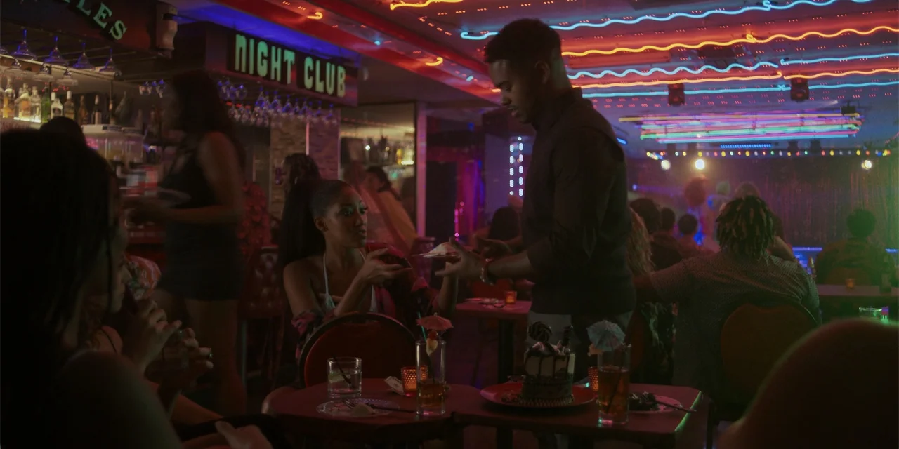Sitting in Bars with Cake (2023) movie still: medium — A dimly lit nightclub features neon ceiling lights and patrons seated at small tables. A man…