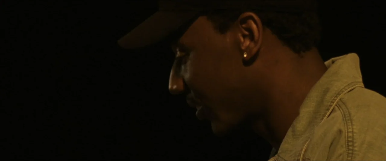 Sermon on the Mount (2019) movie still: closeup — A man stands in a dark room with minimal illumination on his profile. He wears a baseball cap and…