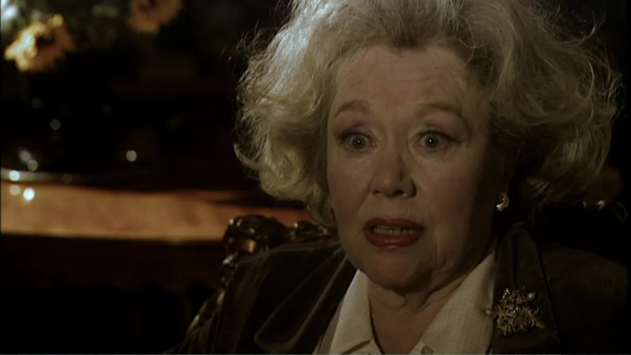 Six by Sondheim (2013) movie still: closeup — An interior space features a dimly lit room with ornate wooden furniture. An elderly woman sits in a…