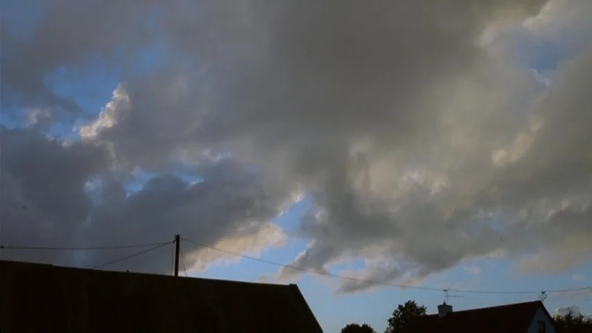 Garage (2007) movie still: establishing — The sky is filled with thick gray clouds interspersed with patches of blue. Dark silhouettes of rooftops…