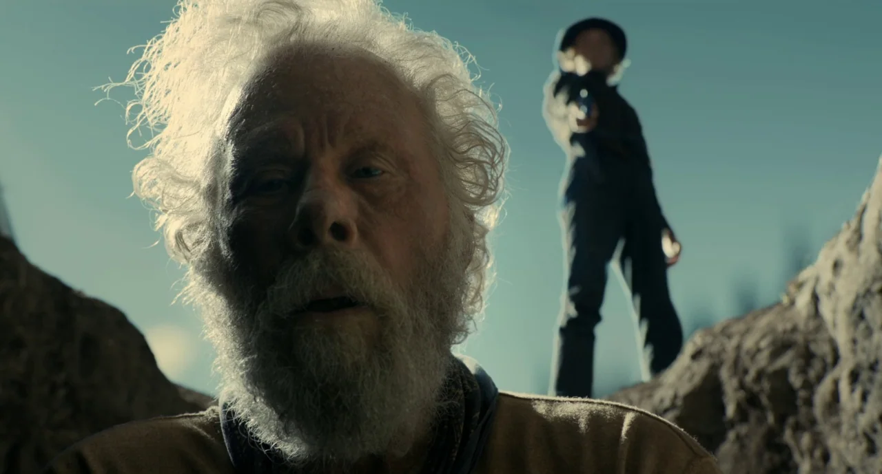 The Ballad of Buster Scruggs (2018) movie still: closeup — A low-angle shot from inside a pit showing an elderly man looking upward. A second figure…