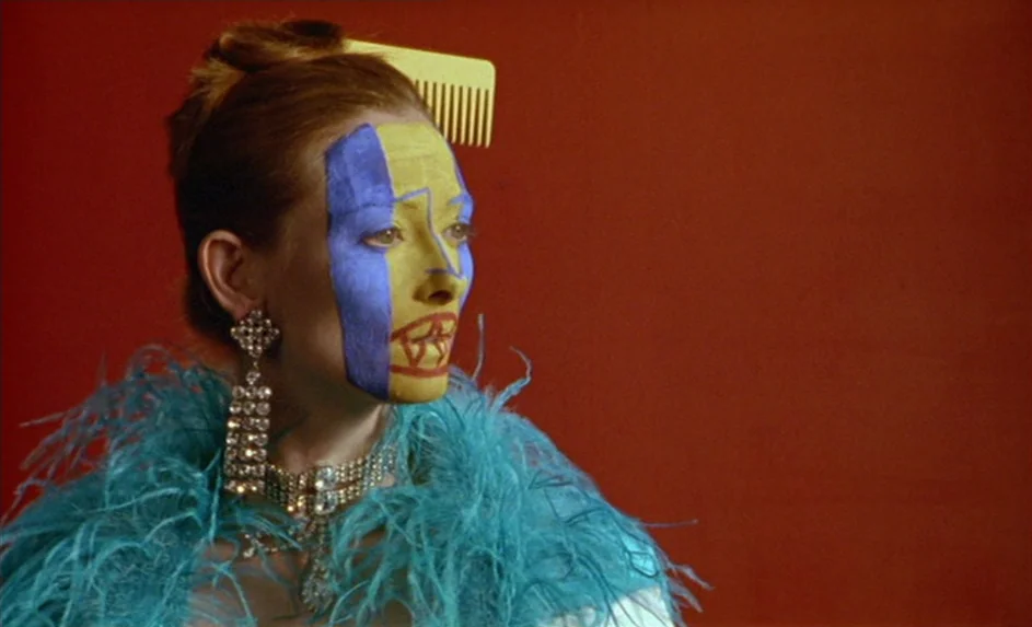 Wittgenstein (1993) movie still: closeup — A woman stands against a solid red wall wearing elaborate jewelry and colorful makeup. She wears a blue…