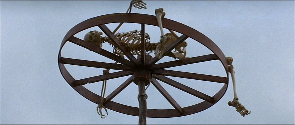 The Devils (1971) movie still: fullbody — A human skeleton is lashed to a large wooden wheel mounted on a pole. The structure is set against a…