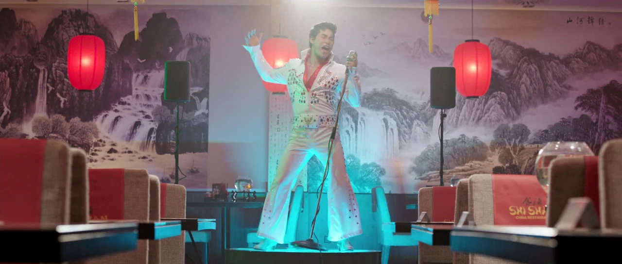 The Voices (2014) movie still: fullbody — A Chinese restaurant interior features a mural of mountains and waterfalls on the back wall. An Elvis…
