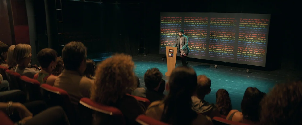 A Hipster in Rural Spain (2024) movie still: wide — An auditorium features a dark stage with a large text display and a speaker. A man stands at a…
