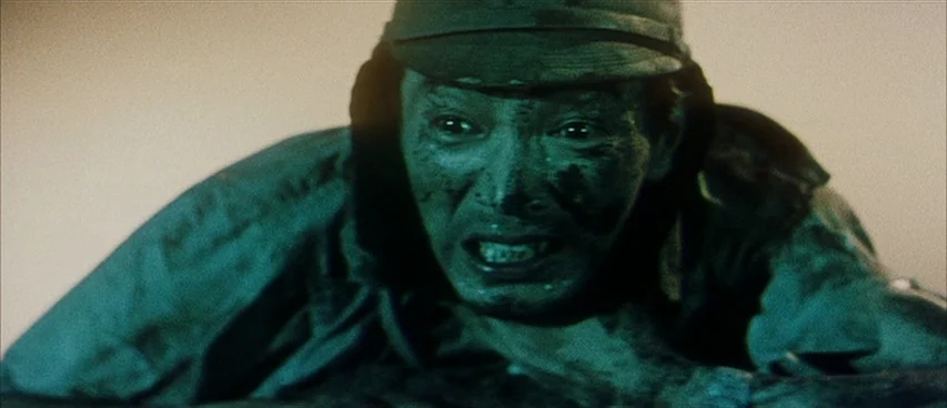 Jigoku (1960) movie still: closeup — A soldier covered in dirt lies close to the ground on a battlefield. The man peers forward with a strained…