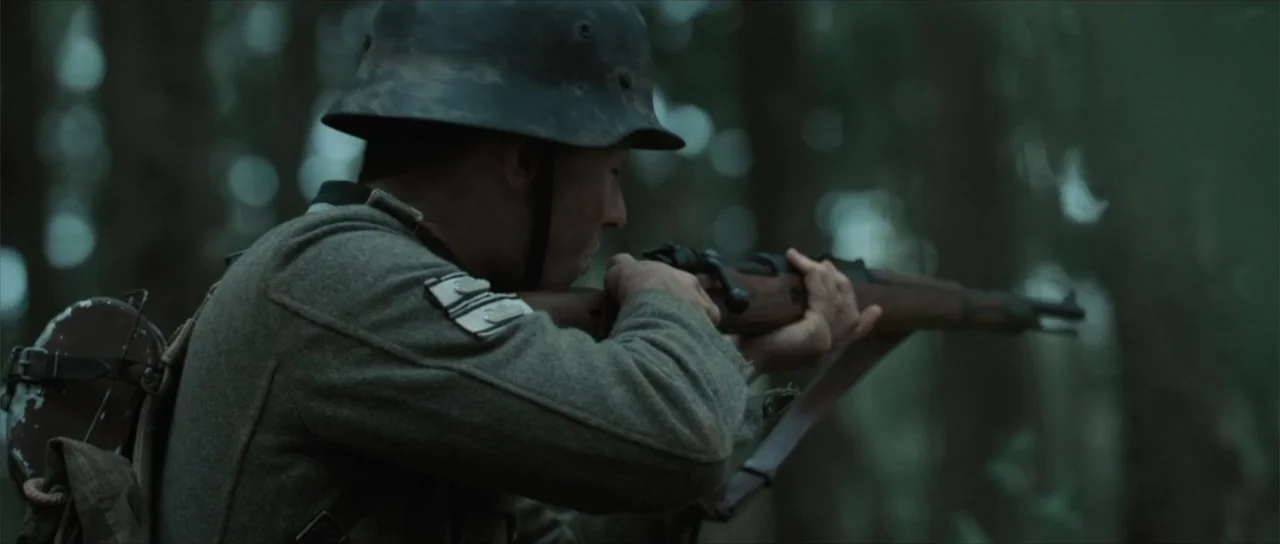 Sin retorno (2009) movie still: medium — A dense woodland environment with blurred tree trunks in the background under daylight. A lone soldier in…