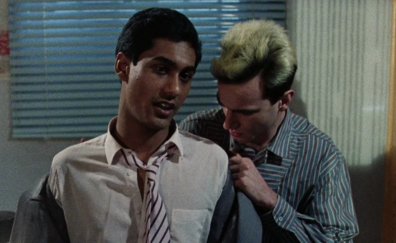 My Beautiful Laundrette (1985) movie still: medium — A small indoor space features a window covered with blue horizontal blinds. Two young men stand…