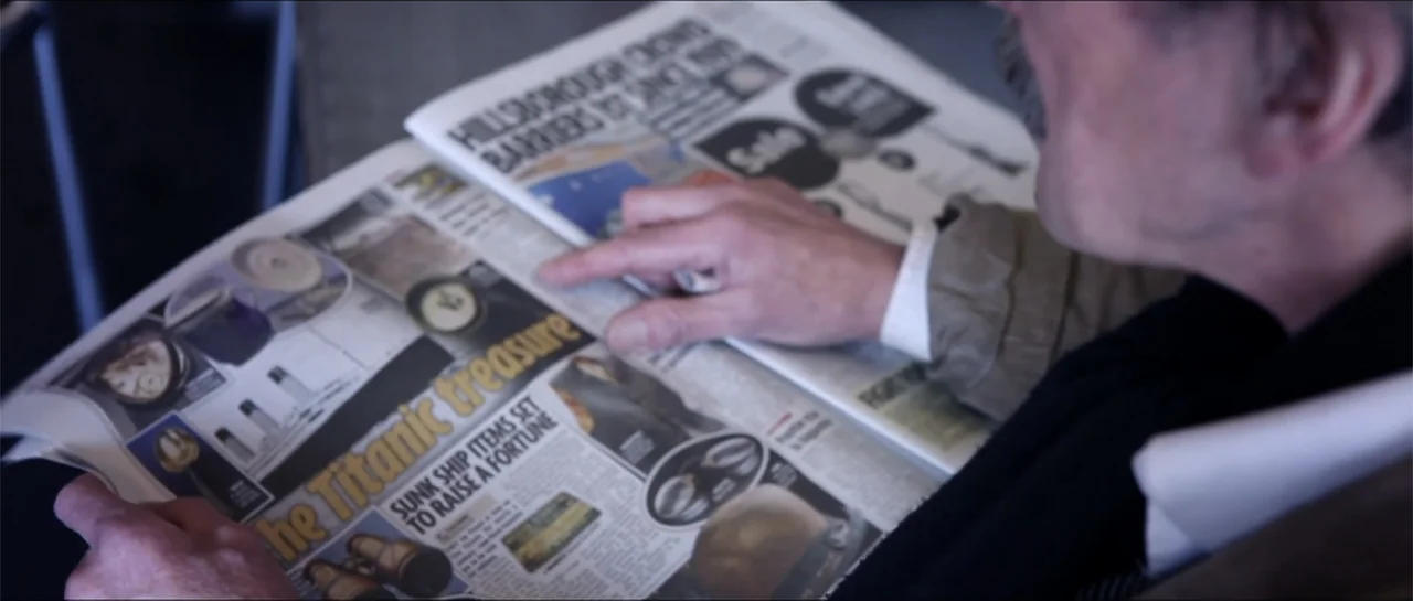 Garnet’s Gold (2014) movie still: closeup — A man holds an open newspaper featuring an article about Titanic artifacts. His finger rests on the page…