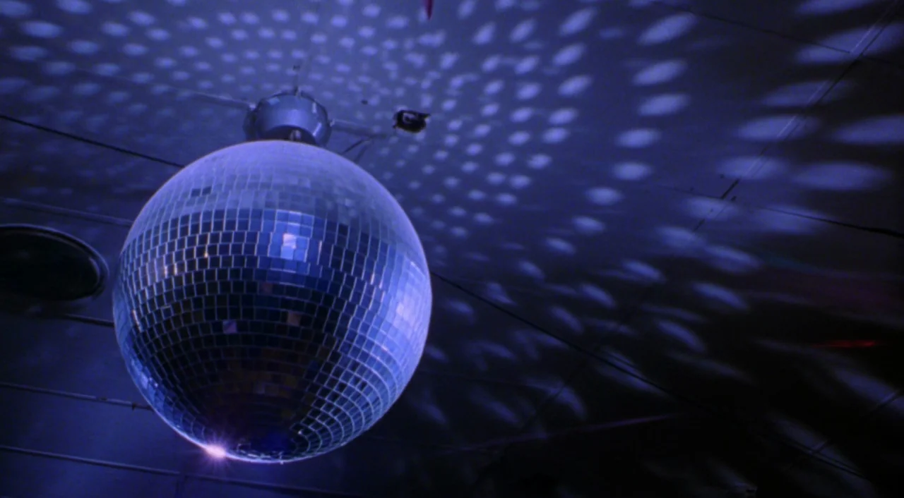 Boys Don’t Cry (1999) movie still: closeup — A spinning mirrored ball is mounted on a dark, panelled ceiling surface. Blue light reflections scatter…