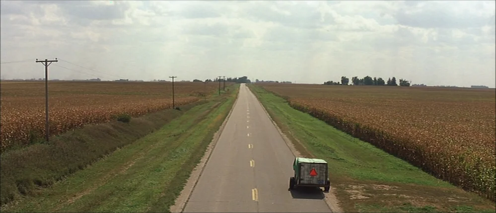 The Straight Story (1999) movie still: wide — A long, straight asphalt road bisects two sprawling, dry cornfields under a cloudy sky. A small green…
