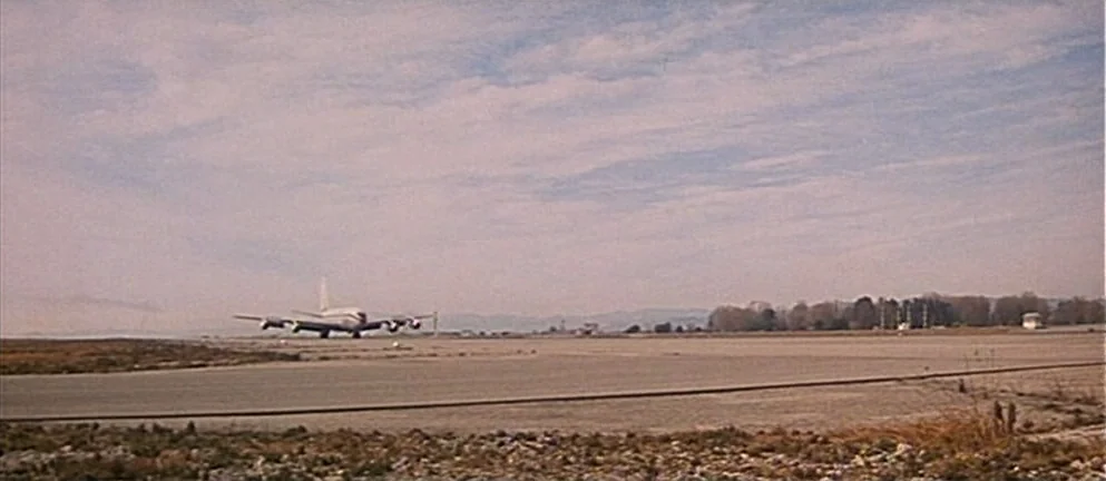 Mississippi Mermaid (1969) movie still: establishing — A vast, flat airfield stretches toward a treeline under a large, overcast, pale sky. An…