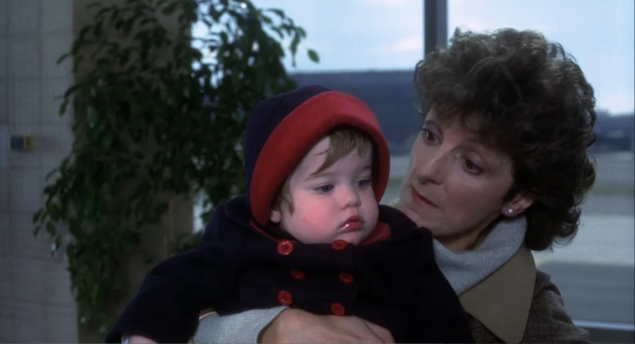 Baby Boom (1987) movie still: medium — An interior space with a large window and a leafy potted plant visible. A woman holds a young child in her…