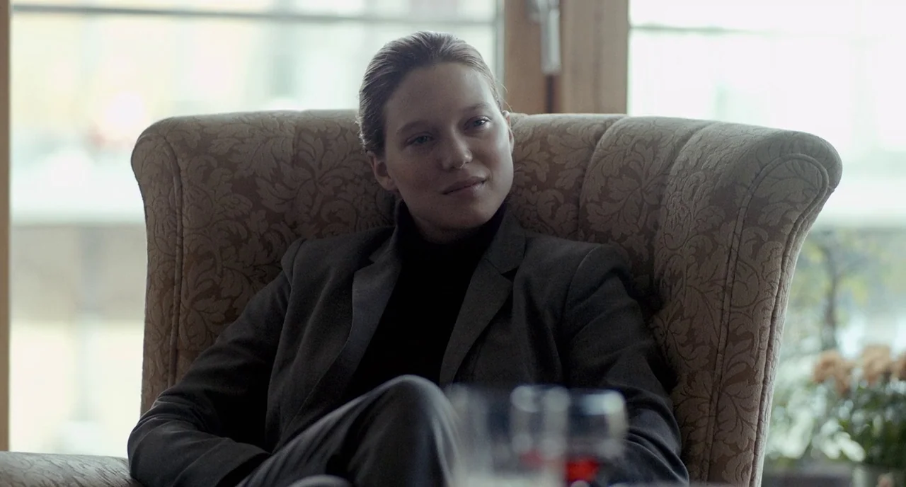 The Lobster (2015) movie still: medium — A woman sits in a patterned upholstered armchair placed before a large window. She wears a grey suit jacket…