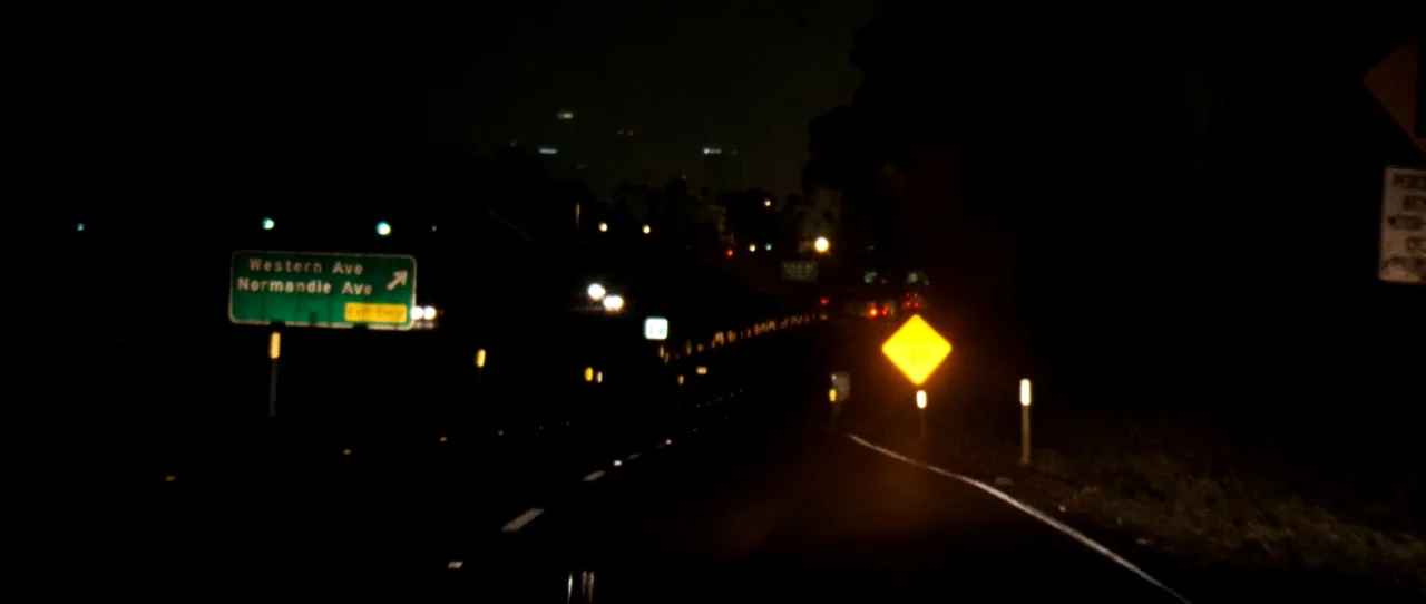 Little Birds (2011) movie still: establishing — A dark night view of a highway exit ramp with reflective green and yellow signage. The road…