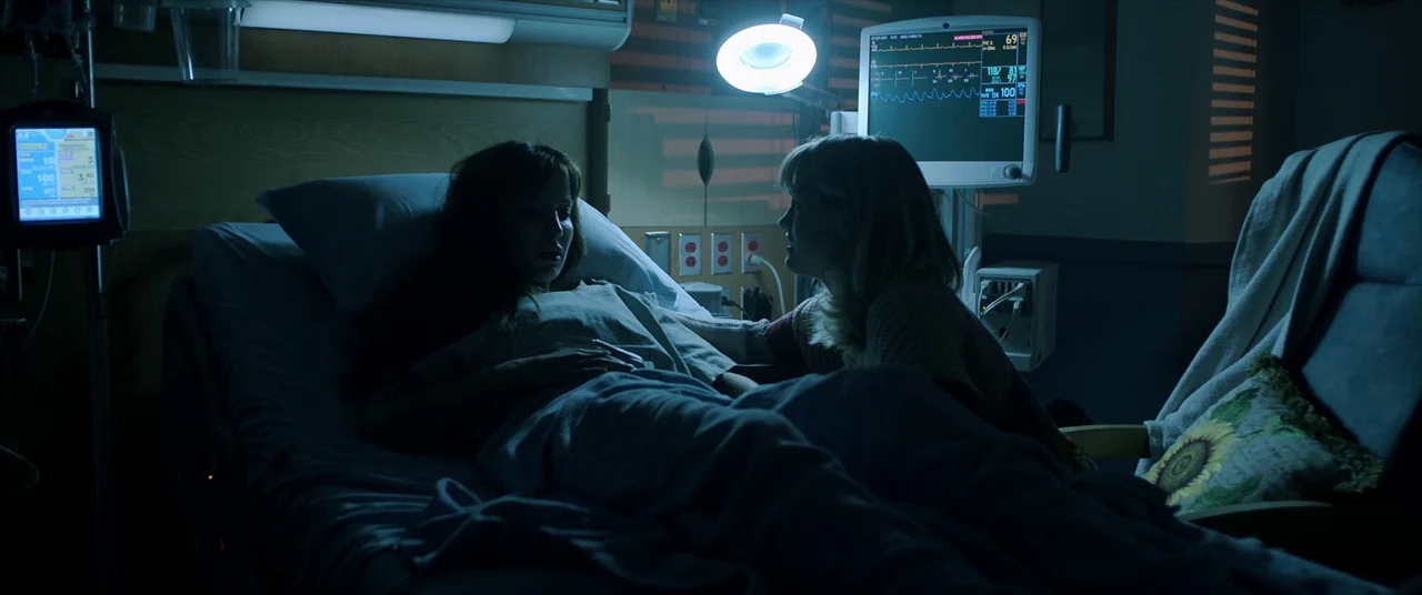 Malignant (2021) movie still: medium — A dark hospital room contains a patient lying in bed beside a bedside visitor. A medical monitor displays…
